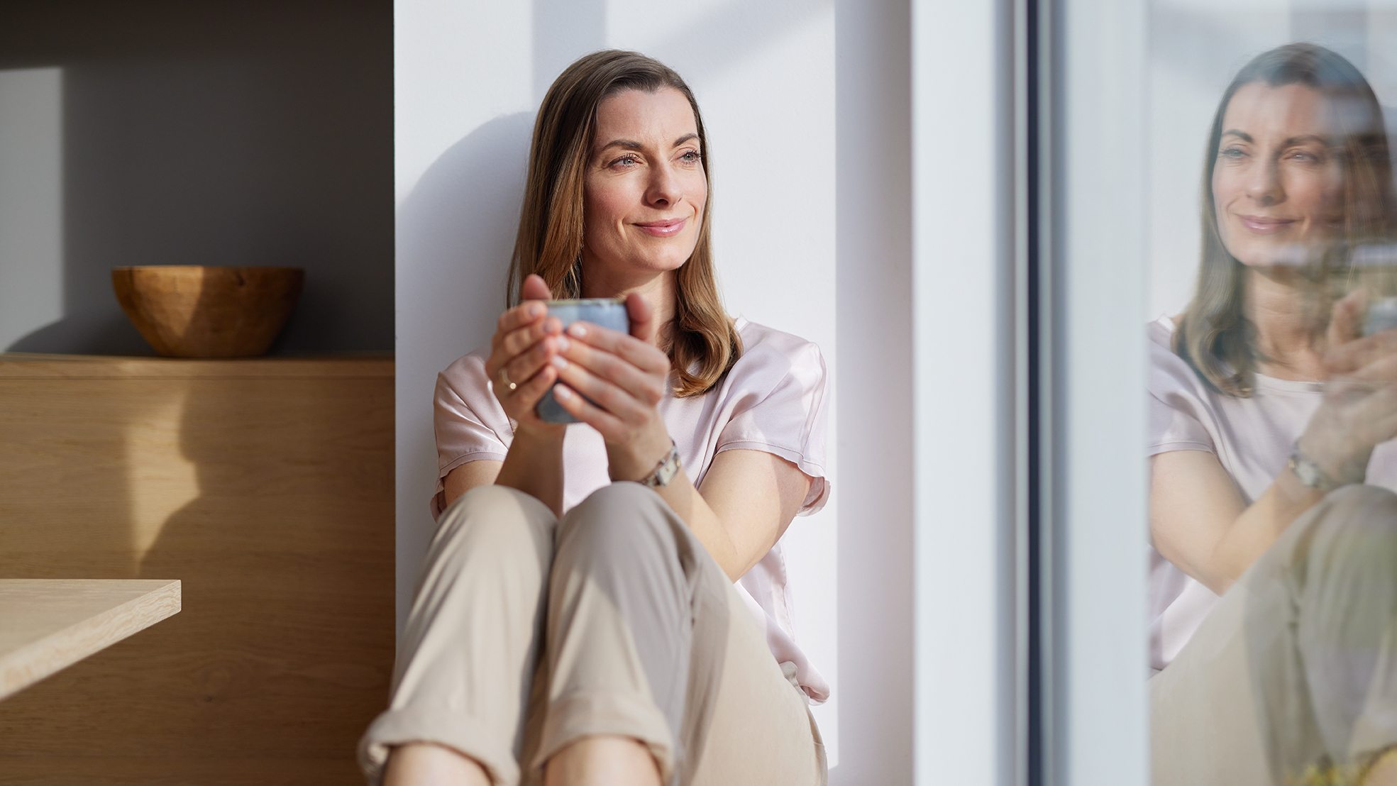 Frau sitzt auf der Fensterbank mit einer Tasse und schaut aus dem Fenster in welchem sie sich spiegelt.